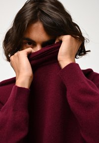 Maglione a coste color bordeaux con un collo alto, caratterizzato da dettagli a coste su polsini e collo, texture morbida, parzialmente oscurato dalle mani.