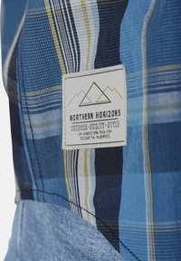 Blaues Karostoff mit einem genähten weißen Etikett, das Bergumrisse und den Text "Northern Horizons Outdoor Utility Style" zeigt.
