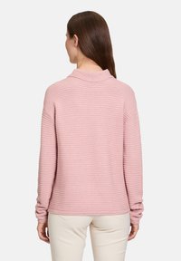 Rosa gerippter Pullover mit hohem Kragen und langen Ärmeln, aus strukturiertem Material und in lässiger Passform, getragen über hellen Hosen.