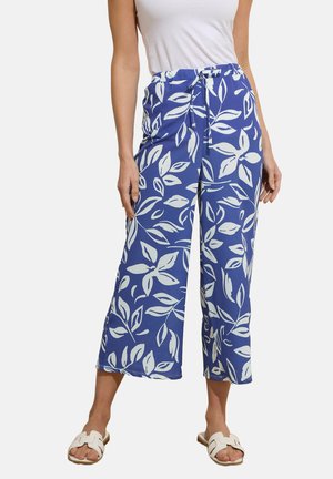 Donna che indossa pantaloni blu a gamba larga e corti con motivi a foglia bianchi e sandali bianchi scivolati, in piedi contro uno sfondo semplice.