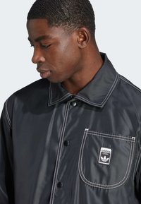 Černá nepromokavá bunda s bílým prošíváním, zapínáním na knoflíky vpředu a náprsní kapsou s logem Adidas. Hladká textura, límcový design.