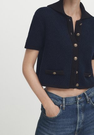 Gros plan d'une personne portant un cardigan bleu marine à manches courtes boutonné avec des bordures noires et un jean bleu taille haute, les mains dans les poches.