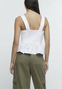 Bílý bezrukávový top s peplum střihem a řaseným zadním dílem, vyznačující se očkovými výřezy podél lemování. V kombinaci s olivovými cargo kalhotami.