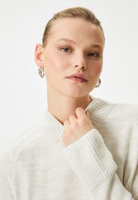 Jasno beżowy sweter w dzianinę z wysokim kołnierzem, prążkowanymi brzegami i teksturowanym wykończeniem, w towarzystwie metalowych kolczyków w kształcie kół.