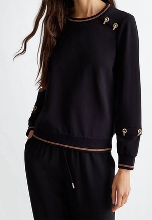 Kobieta ubrana w czarną bluzkę z długim rękawem z brązową lamówką i ozdobnymi złotymi nacięciami na dekolcie i mankietach, w połączeniu z czarnymi spodniami.