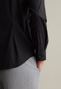 Schwarzes Hemd mit langen Ärmeln, Knopfleiste, abgerundetem Saum und geknöpften Manschetten. Kombiniert mit hellgrauen, fein gestreiften Hosen.