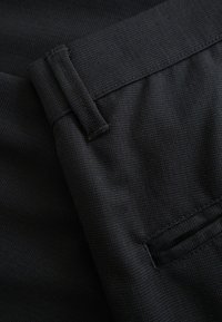 Közelkép fekete texturált szövetnadrágról, fókuszban a derékpánt, az övbújtató és a varrott hátsó zseb részlete.