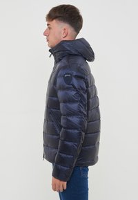 Giacca puffer blu navy con finitura lucida, cappuccio, tasche con zip e pannelli cuciti. Presenta un logo stampato sulla spalla sinistra.