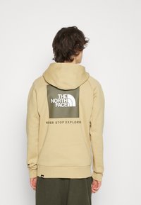 Felpa beige con cappuccio, dotata di un quadrato verde scuro sul retro con testo e logo bianchi. Polsini e fondo a costine, morbida texture.