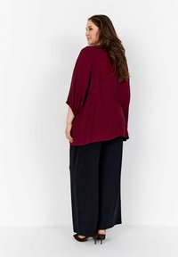 Blusa borgogna con vestibilità ampia e maniche a tre quarti, abbinata a pantaloni neri a gamba larga e scarpe con tacco basso. Tessuto dalla texture liscia.