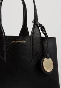 Svart läder tote-väska med pebble-textur, med en guldfärgad logotyp, en fäst cirkulär charm och korta handtag med vikta detaljer.