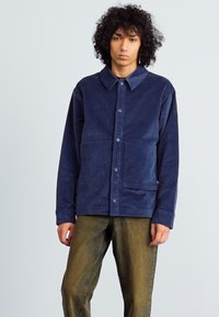 Navy blue jacket made of soft fabric with a collar, front buttons, and two large pockets; worn with olive-green pants.