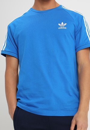 Mężczyzna w jaskrawo niebieskiej koszulce Adidas z białymi paskami na rękawach i logo Adidas na piersi, stoi z rozluźnionymi rękami.