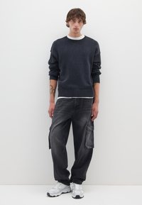 Donkergrijze gebreide trui met geribbelde manchetten, gecombineerd met loszittende donkere cargobroek met zijzakken. Model draagt witte sneakers.