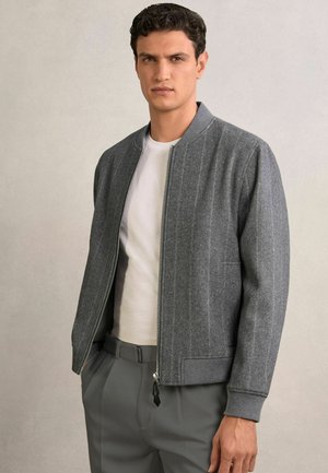 Tânăr purtând o jachetă cu fermoar de culoare gri deschis peste o cămașă albă și pantaloni gri închis, stând pe un fundal simplu de culoare deschisă.