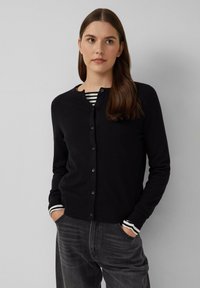 Cardigan noir avec un col rond et une fermeture à boutons. Porté sur une chemise rayée noire et blanche. Le matériau semble doux et texturé.