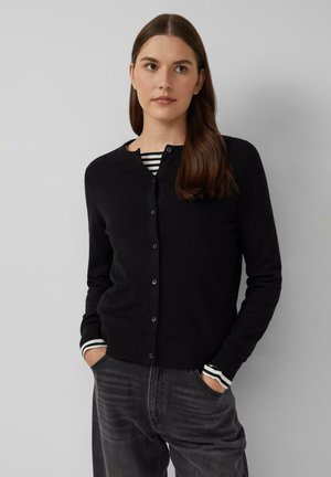 Cardigan noir avec un col rond et une fermeture à boutons. Porté sur une chemise rayée noire et blanche. Le matériau semble doux et texturé.
