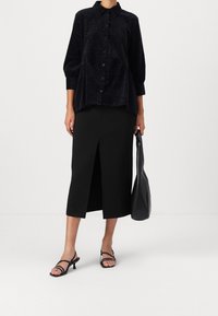 Femme portant un chemisier noir à boutons, une jupe midi noire avec une fente à l'avant, des sandales noires, tenant un sac à main en cuir noir à ses côtés.