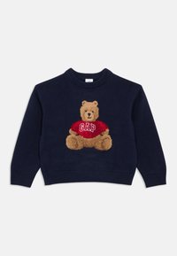 Tumši zila adīta džemperis ar tāfeles (teddy bear) grafiku, kas valkā sarkanu krekliņu ar "GAP" tekstu, apaļu dekoltē un ribotām piedurknēm un apakšmalu.