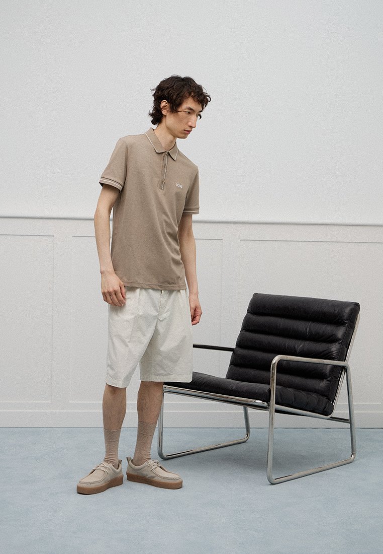 Hellbraunes Poloshirt mit Kragen, weiße Shorts, beige Turnschuhe und graue Socken. Steht neben einem modernen schwarzen Lounge-Stuhl.