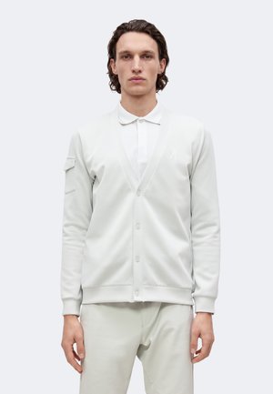 Homme de face portant un polo blanc, un cardigan clair boutonné et un pantalon clair assorti sur un fond uni clair.