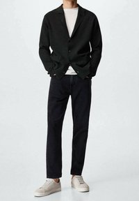 Blazer nero intrecciato su una t-shirt bianca; jeans scuri; sneakers beige. Il blazer presenta un colletto a rever e due bottoni.