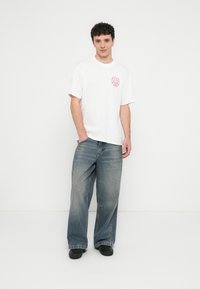 T-shirt branca de algodão com um desenho gráfico vermelho no bolso do peito esquerdo, combinada com jeans azuis largos e sapatos pretos.