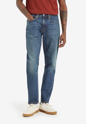 Uomo che indossa jeans blu, sneakers bianche e una maglietta color ruggine, in piedi con una mano in tasca davanti a uno sfondo uniforme.