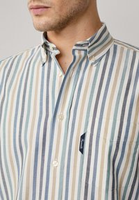 Camisa de botones a rayas con líneas verticales en azul suave, beige y blanco. Material de algodón con un bolsillo en el pecho y etiqueta de logo.