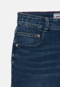 Jeans de mezclilla azul oscuro con un diseño clásico, que cuentan con cinco bolsillos, herrajes dorados y detalles de costura visibles. Textura suave.