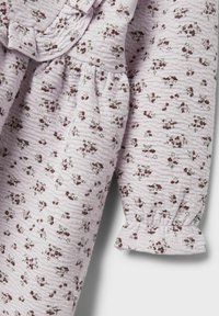 Ljust lila tyg med små blommönster i mörkröd och grön. Har en volanghalsringning och elastiska ärmslut. Texturerad yta.