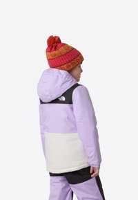 Lilla og svart vinterjakke med en hvit nedre del; brukes med en rosa og gul mønstret strikkelue med dusk.