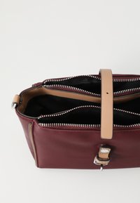 Geantă de mână din piele burgundă, cu două compartimente cu fermoar, curea din piele bej și accente metalice argintii. Textură netedă cu detalii cusute.