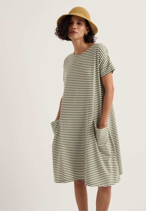 Vrouw draagt een loszittende, knie-lange jurk met groene en witte strepen, korte mouwen en grote voorzakken, gecombineerd met een geweven hoed.