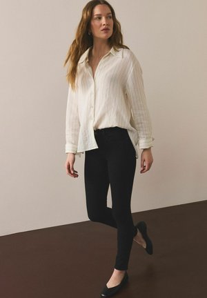 Noor naine kõndimas siseruumides, kandes lahtist kreemika triibulise särgiga, musti kitsaid pükse ja musti madalaid kingi, taust on lihtne.