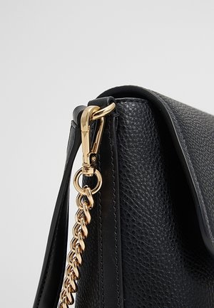 Róg czarnej torebki ze skóry o fakturze, z detalem złotego łańcuszka i zapięcia na pasku, na jasnoszarym tle.