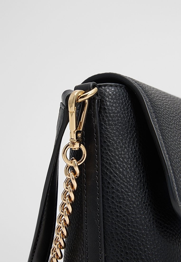 Coin d'un sac à main en cuir noir texturé avec une chaîne dorée et un détail de fermoir sur la sangle, sur un fond gris clair.
