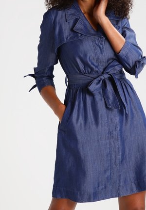 Femme portant une robe en jean ceinturée, longueur genou, avec des manches 3/4 nouées aux poignets et la main dans la poche.