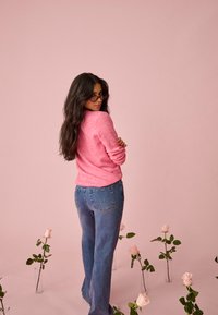 Kvinde med langt mørkt hår, lyserød sweater og briller står med krydsede arme blandt lyserøde roser i glasvaser på en blød lyserød baggrund.