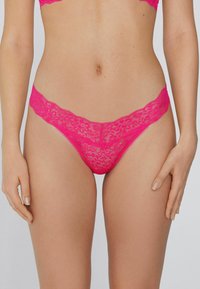 String en dentelle rose vif avec un motif floral, des bords festonnés et une texture délicate, offrant une couverture minimale et une finition douce.