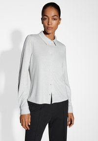 Camicia grigia chiaro a maniche lunghe con colletto, realizzata in un tessuto strutturato, con bottoni frontalmente e maniche con polsino. Indossata con pantaloni scuri.