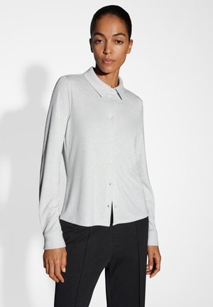 Camicia grigia chiaro a maniche lunghe con colletto, realizzata in un tessuto strutturato, con bottoni frontalmente e maniche con polsino. Indossata con pantaloni scuri.