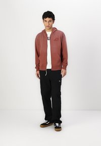 Ruda zip-up gobtuvė su gobtuvu ir priekinėmis kišenėmis, dėvima ant baltos marškinėlių. Sujungta su juodais kargo džinsais ir juodais sportbačiais.