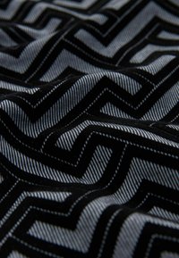 Melnas un pelēkas audums ar zigzag rakstu. Tekstūra ir mājīga, ar pretstatā esošām gludām svītrām, kas rada dimensiju efektu.