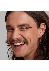 Porträt eines Mannes mit langen braunen Haaren und einem hellen Schnurrbart, der Ohringe und kräftigen Eyeliner trägt und vor einem neutralen Hintergrund lächelt.