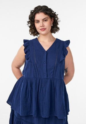 Mladá žena s krátkymi kučeravými vlasmi, ktorá má na sebe tmavomodrú blúzku bez rukávov s volánikmi, stojaca na bielom jednoduchom pozadí.
