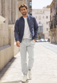 Marine bomberjack in laagjes over een wit T-shirt, gecombineerd met lichtgrijze joggingsbroek en witte sneakers, wandelend langs een straat met stenen klinkers.
