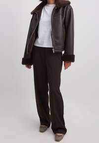 Veste en cuir marron avec un col et des manchettes en fourrure brune, associée à un t-shirt blanc et un pantalon marron. Des chaussures plates marron complètent le look.