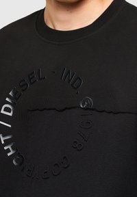 Černá bavlněná mikina s texturovaným písmem a kruhovým designem na hrudi. Hladký výstřih a výrazné detaily z látky.