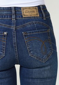 Jeans de mezclilla azul oscuro con una etiqueta de cuero en la parte posterior. Presenta dos bolsillos traseros con costuras amarillas y un diseño de costura decorativa.